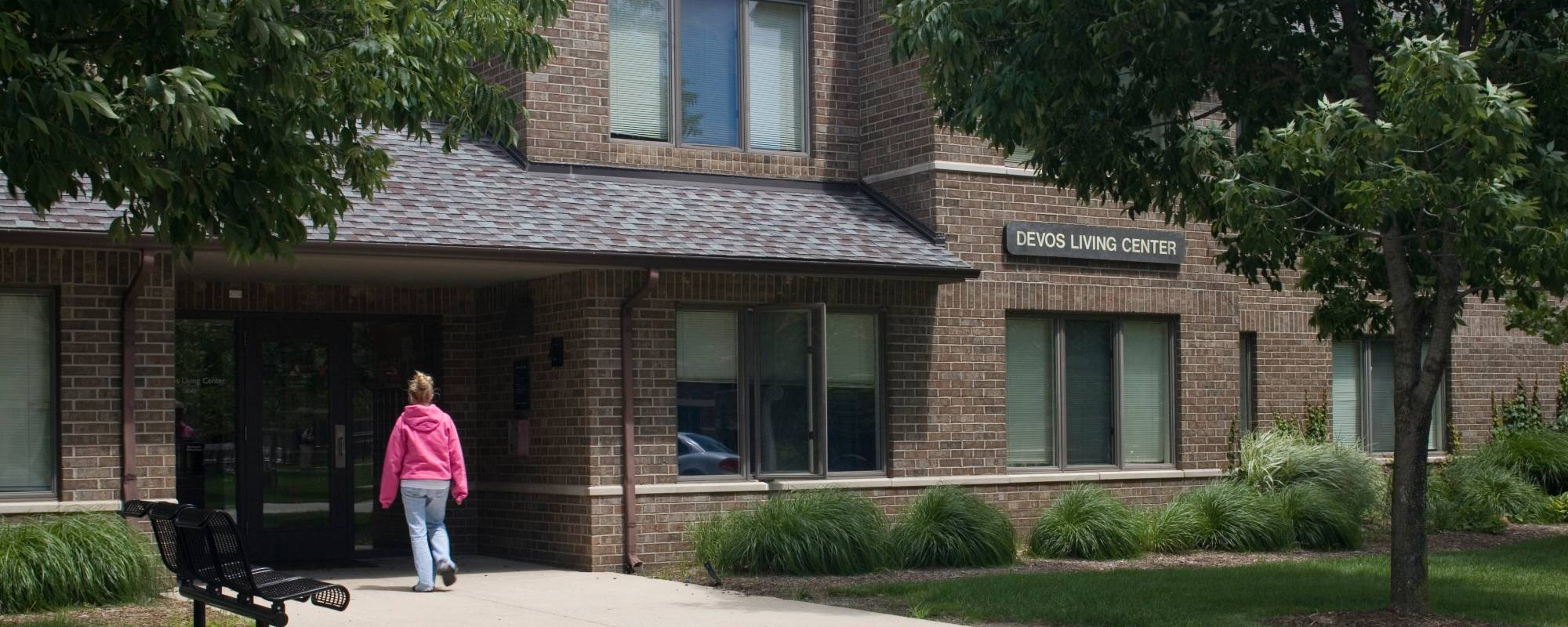 A person in a pink hoodie walks toward a brick building labeled "DeVos Living Center" surrounded by green shrubs and trees, creating a welcoming atmosphere.